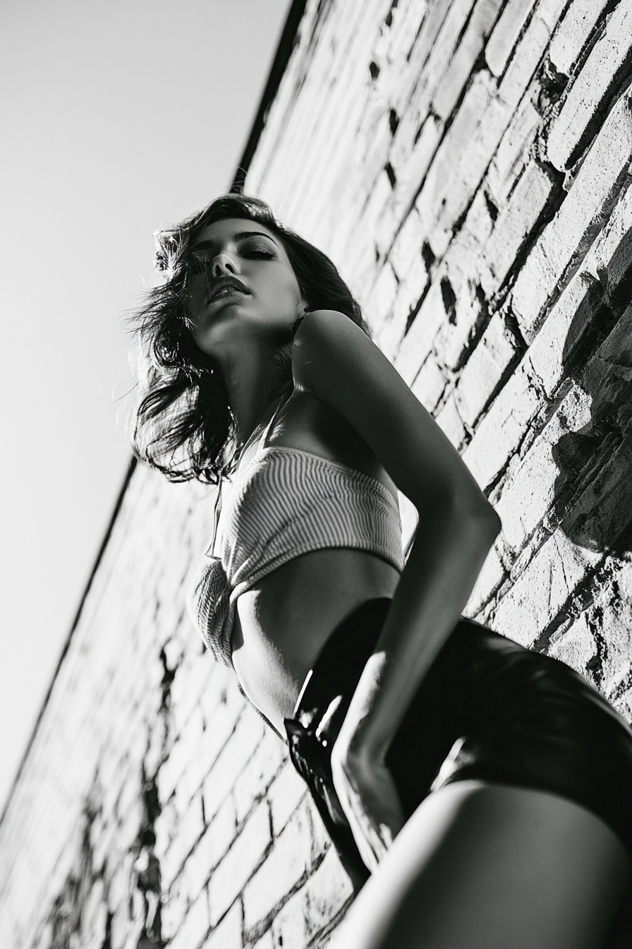 Black and white urban fashion portrait of a woman leaning against a brick wall in dramatic low-angle photography