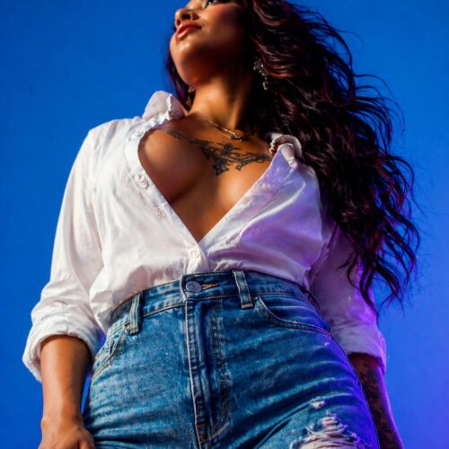 A powerful portrait of a woman with long dark hair and intricate chest and thigh tattoos, looking upward with a confident expression against a blue and purple light backdrop