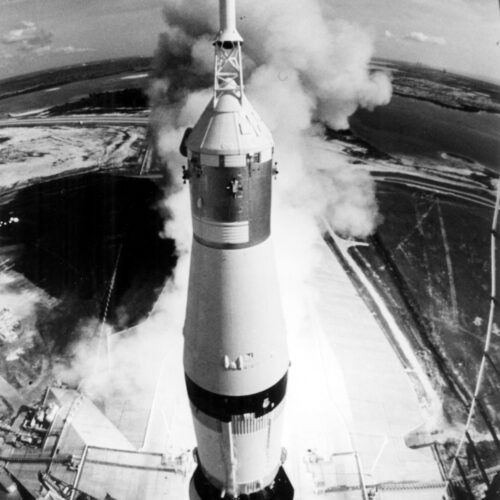 High-angle black and white photographic poster of a space rocket launching from a pad with smoke and flame.