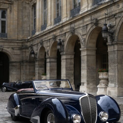 Photographic poster of a glossy midnight-blue vintage Bugatti convertible parked in an elegant Parisian courtyard with classical architecture and arched colonnades.