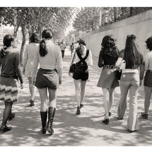 Black-and-white vintage photograph of stylish women walking down a sun-dappled urban street with trees and buildings, evoking mid-century street fashion.