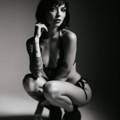 Black and white intimate portrait of a sensual tattooed woman in contemplative pose with dramatic lighting and high contrast monochrome photography.