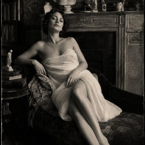 Vintage noir portrait of a contemplative woman in draped fabric, seated in an ornate interior with classical busts and period furnishings, monochromatic photography.