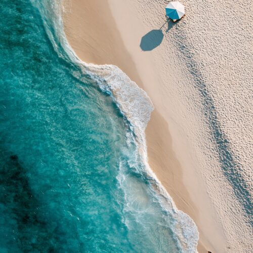Aerial photographic view of a serene beach with turquoise water, white sand, and a figure with blue umbrella casting a shadow on pristine shore.