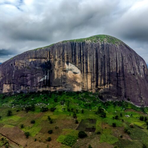 Photographic aerial view of Zuma Rock in Niger State, Nigeria, showing distinctive columnar basalt formations with green savanna landscape below and cloudy sky.