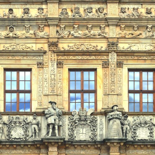 Ornate Renaissance facade of Brzeg Castle with sculptural stone details, classical figureheads, and elaborate carved stonework surrounding tall windows.