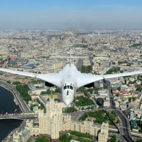 White Tupolev Tu-160 supersonic bomber flying above Moscow cityscape with river, historic architecture, and sprawling urban landscape below.