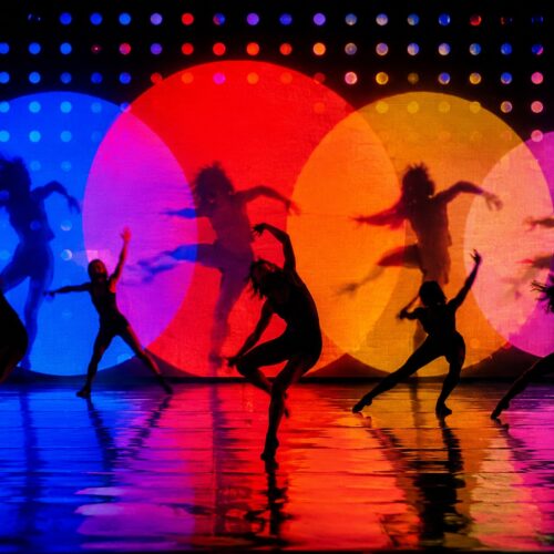 Pop art poster of silhouetted dancers leaping against glowing neon blue, red, orange, and magenta circles with dynamic movement and reflections.