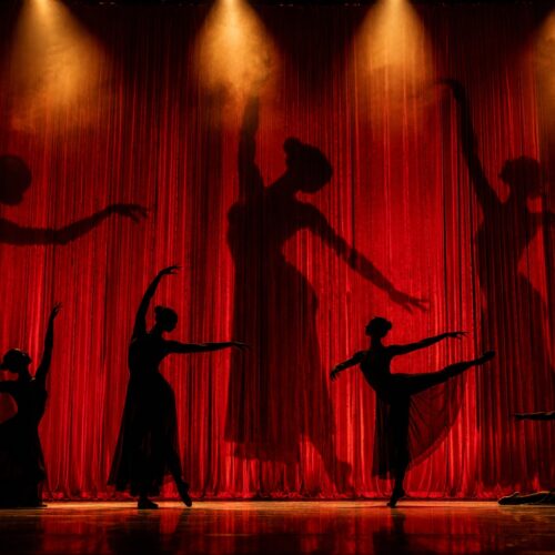 Silhouettes of graceful ballet dancers performing on a red stage curtain with golden spotlight illumination creating dramatic theater wall art.
