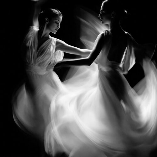 Black and white noir poster of two dancers moving in synchronized harmony with flowing fabrics dissolving into ethereal light against deep black background.