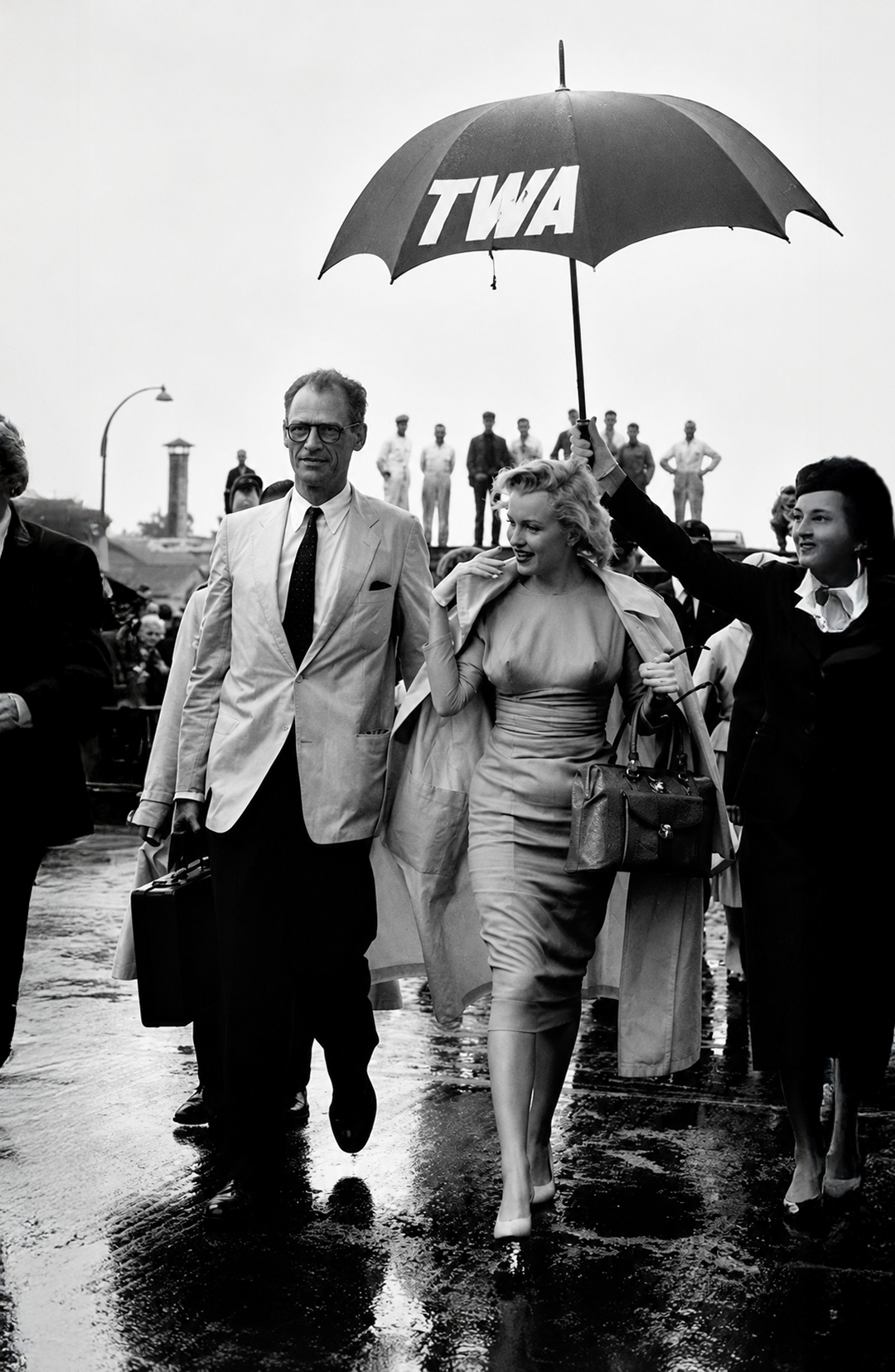Black-and-white vintage photograph of Marilyn Monroe and Arthur Miller walking under a TWA umbrella at London airport in 1956, iconic Hollywood wall art poster.