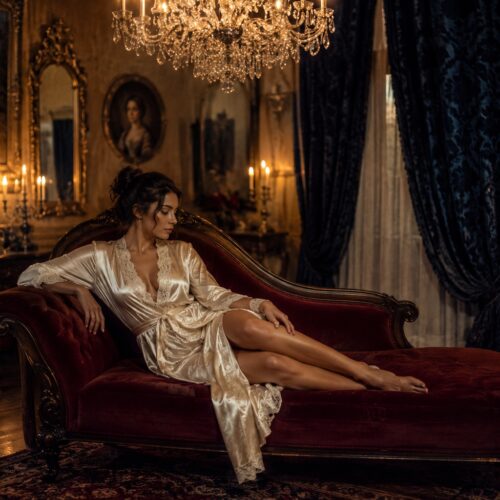 Baroque woman in ivory silk robe reclining on crimson velvet chaise lounge in ornate gold-framed room with crystal chandelier and candlelit luxury wall art.