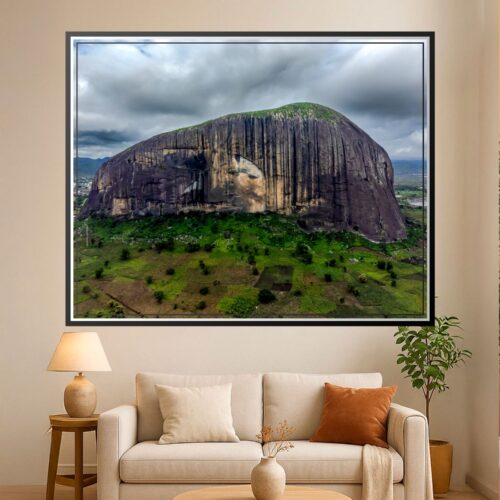 Photographic aerial view of Zuma Rock in Niger State, Nigeria, showing distinctive columnar basalt formations with green savanna landscape below and cloudy sky.